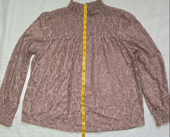 A mauve lace long-sleeve top laid flat on a white fabric backdrop with a yellow tape measure running vertically down the center to show dimensions. A black rolled fabric cover is visible at the bottom.