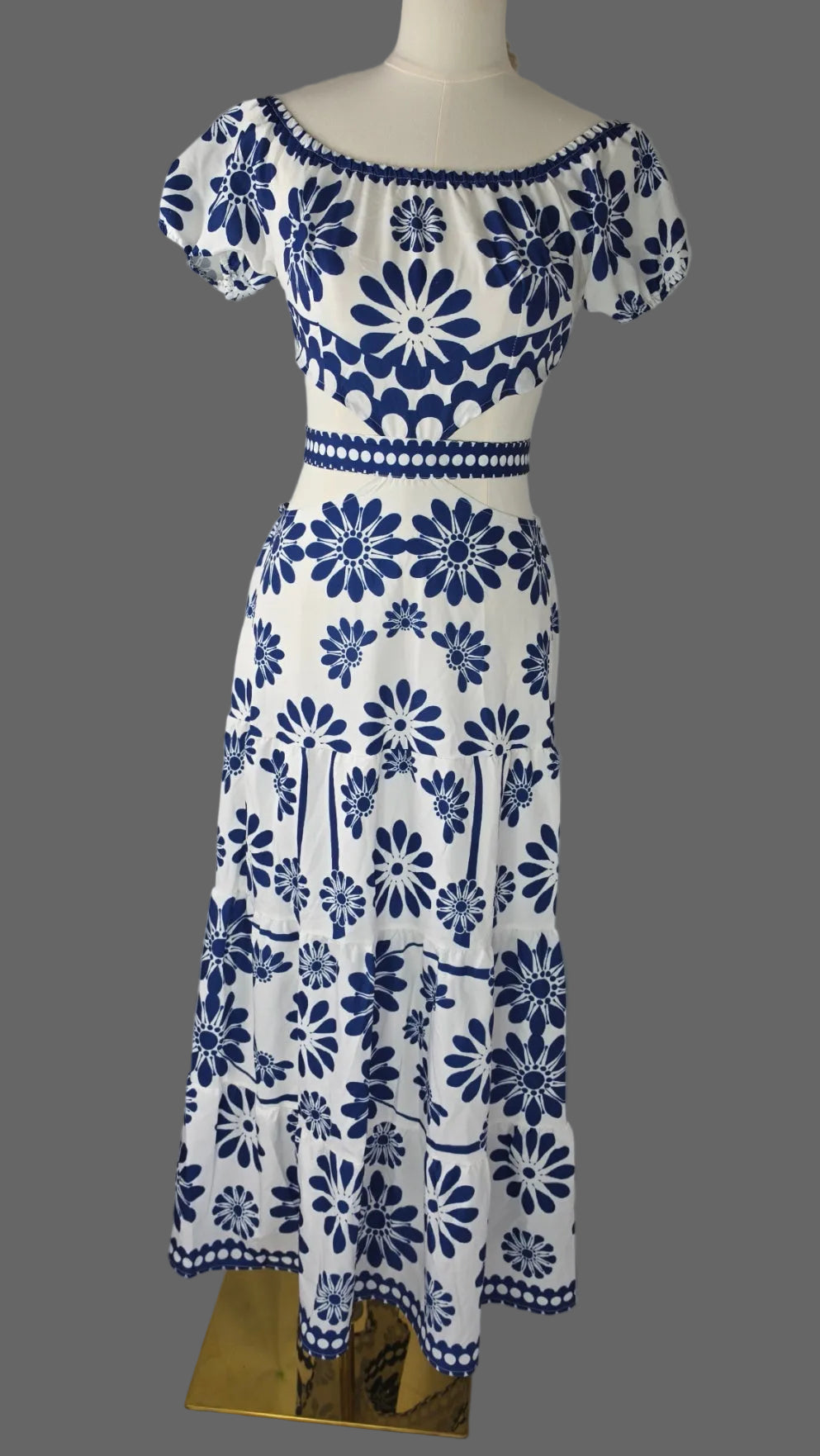 A white off-the-shoulder maxi dress with a blue floral and geometric pattern, displayed on a mannequin against a white studio background.