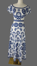 A white off-the-shoulder maxi dress with a blue floral and geometric pattern, displayed on a mannequin against a white studio background.