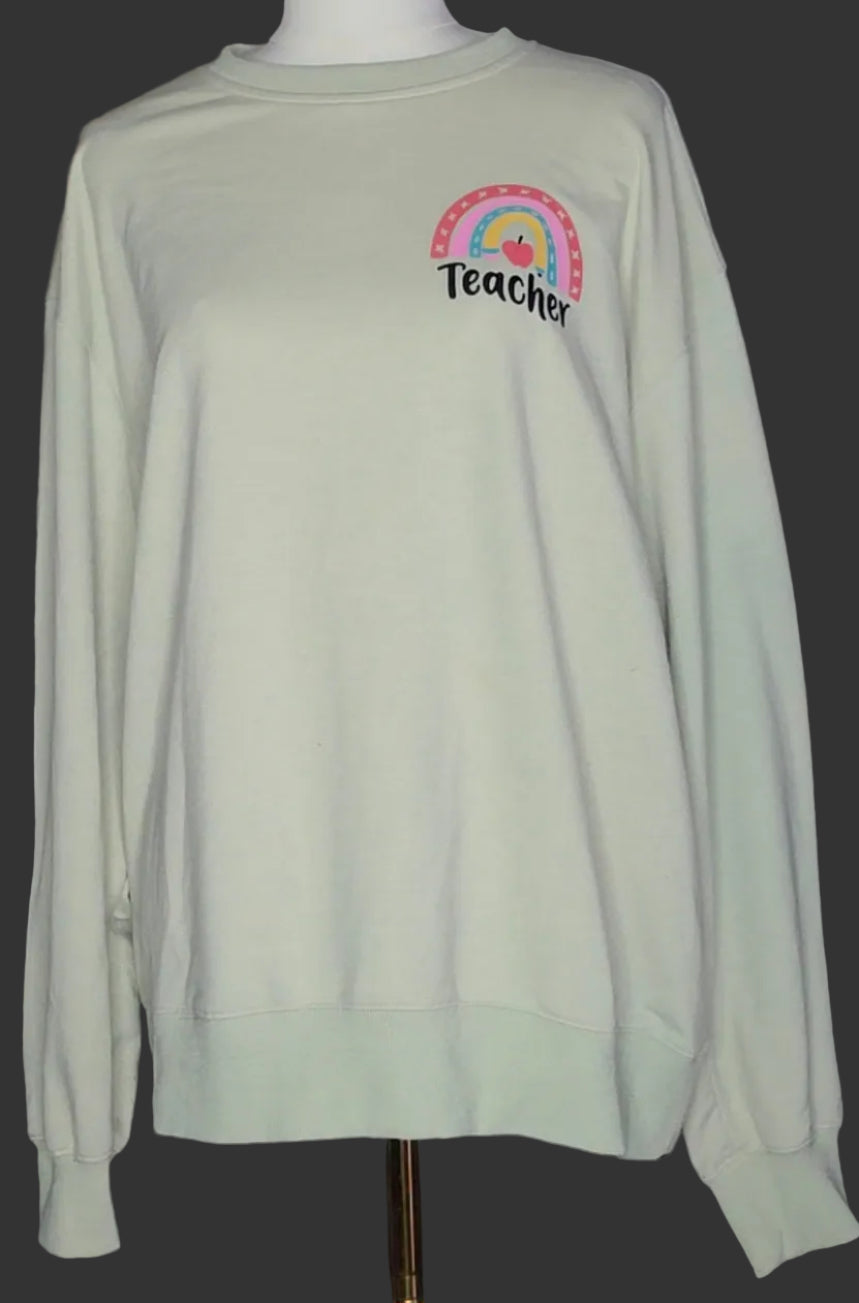 A sage green crewneck sweatshirt with a colorful rainbow and 'Teacher' text graphic on the left chest, displayed on a mannequin. A yellow hoodie hangs on a rack in the background.