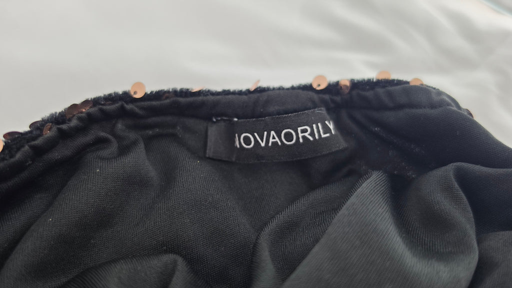 Close-up of a black fabric garment with a visible sewn-in label reading NOVAORILY, showing a gathered elastic waistband with small metallic embellishments.
