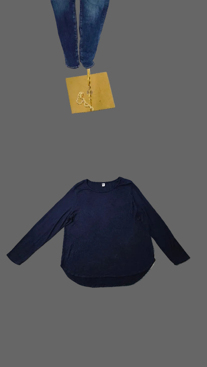 A navy blue long-sleeve crewneck shirt laid flat on a light blue and white patterned rug in a room setting, with a white draped backdrop, carpet, and other items visible in the background.
