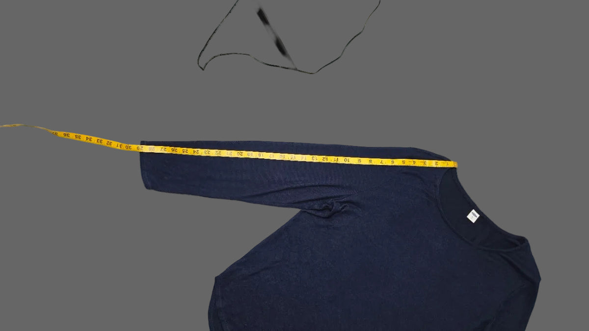 A navy blue long-sleeve shirt laid flat on a light blue and white patterned rug, with a yellow measuring tape stretched across the chest to show the sleeve length. A black tripod leg and a white garment are also visible in the scene.